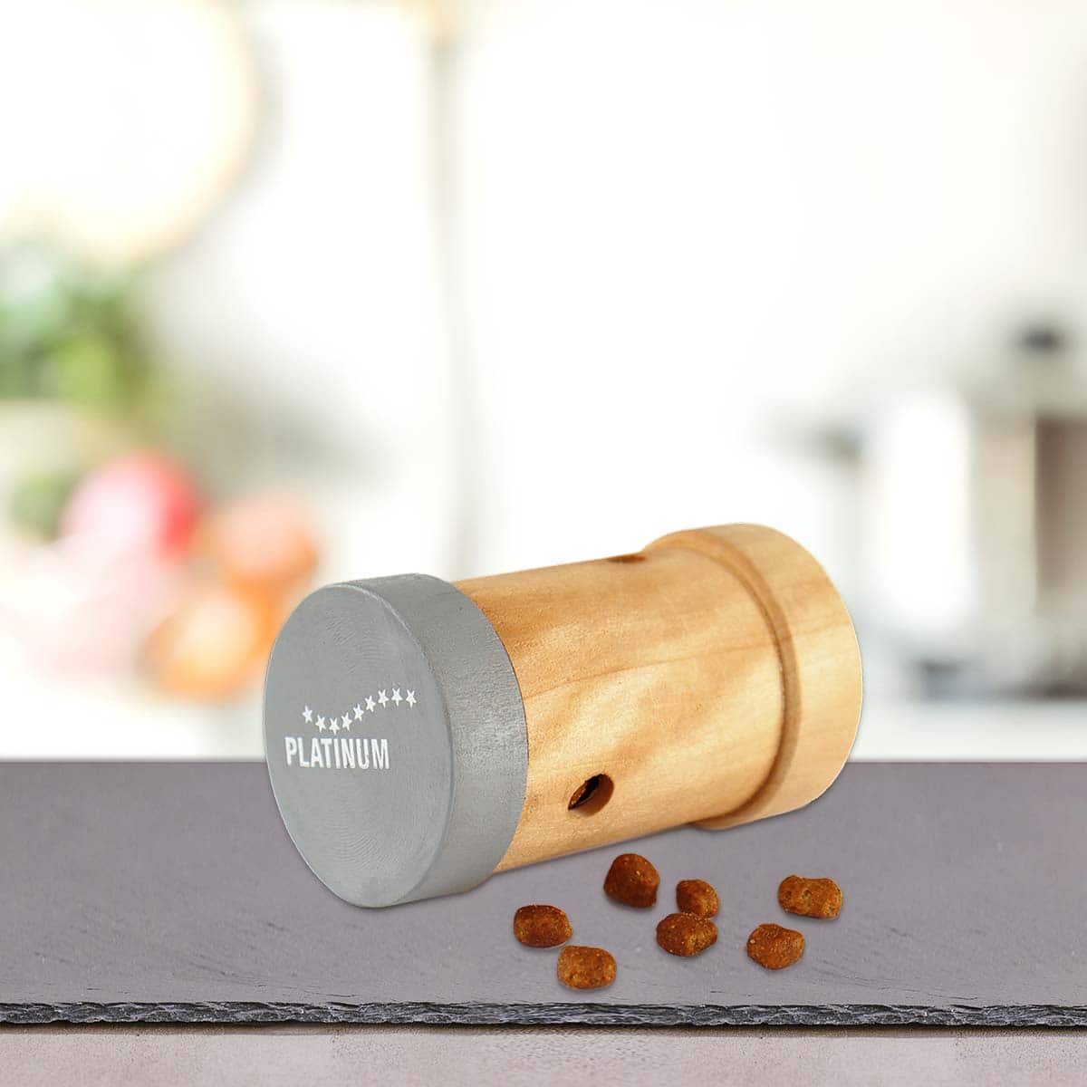 PLATINUM Activity Snack Roll za mačke: Vključujejo: Zabava in nagrada v enem leži na boku na radnoj površini, okružen komadićima hrane, pružajući mentalnu stimulaciju. Pozadina je nježno zamućena.