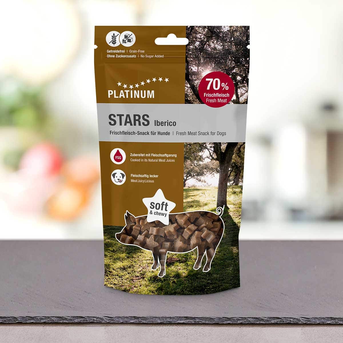Na pultu stoji vrečka PLATINUM Iberico Stars 185 g - Nepremagljiva nagrada iz 100 % svežega mesa Iberico. Na embalaži je silhueta prašiča, napolnjena z dobrotami, v ozadju pa so vidna drevesa.