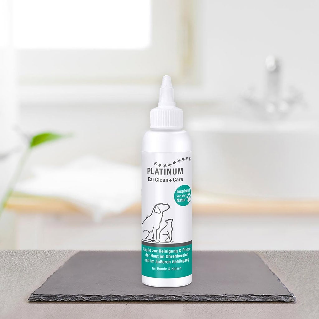 Steklenička izdelka PLATINUM Ear Clean + Care, Nega Ušes za Pse in Mačke, stoji na črni ploščici v svetli kopalnici, v ozadju pa sta zamegljena umivalnik in brisača.