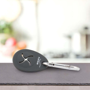 Črn oval Nosilec vrečk za iztrebke by PLATINUM s kovinsko sponko leži na sivi površini, na kateri so napis "PLATINUM" in zvezde v beli barvi. Zamegljeno kuhinjsko ozadje namiguje, da je idealno za sprehod s psom.