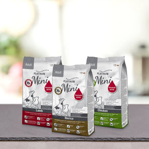 Na pultu so ena poleg druge postavljene tri vrečke PLATINUM Paket različnih okusov Mini Suhe Hrane za Pse, vsaka v pisani embalaži. Ozadje je zamegljeno, kar poudarja vrhunsko blagovno znamko.