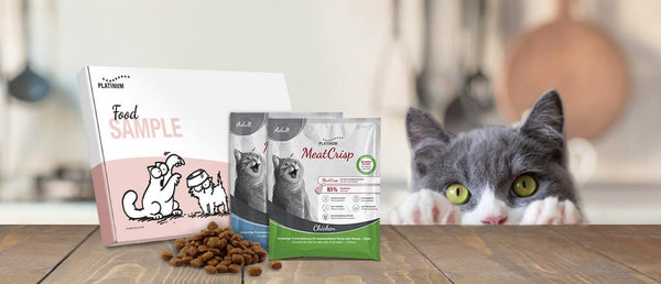 Na mizi so škatla z napisom Vzorec hrane, dve vrečki mačje hrane Platinum in kupček drobovine. Sivo-bela mačka z rumenimi očmi gleda čez rob v ozadju.