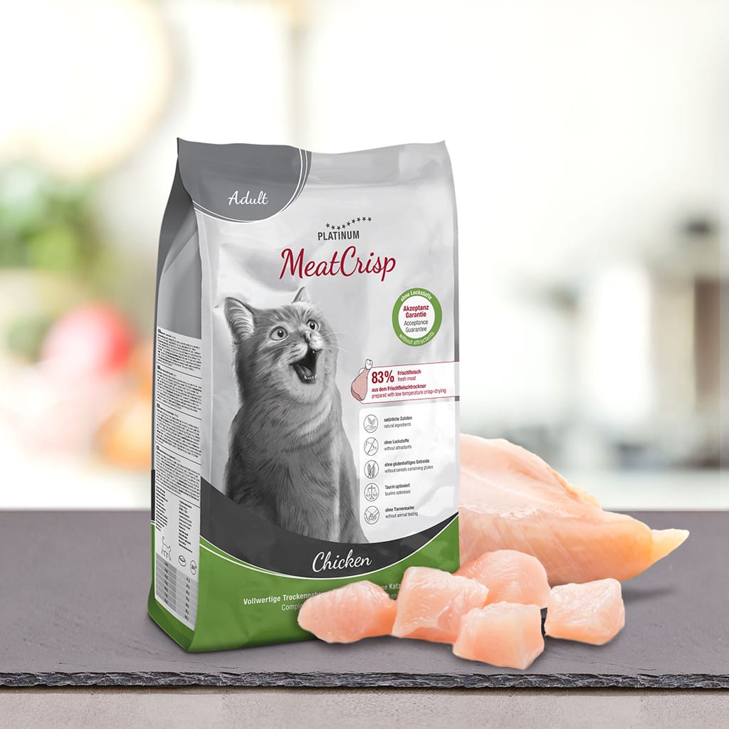 Vrečka PLATINUM MeatCrisp Adult Chicken, Suha Hrana za Mačke, leži na pultu poleg kosov surovega piščanca. Na embalaži so prikazani postopek Freshmeatdryer, poudarjene sestavine in srečna mačka.