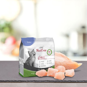 Vrečka PLATINUM MeatCrisp Kitten Chicken, Suha Hrana za Mačke leži na pultu s sivo mačko in kosi surovih piščančjih prsi na ozadju kuhinje, ki je mehko zamegljeno in poudarja postopek sušenja v sušilnici Freshmeatdryer.