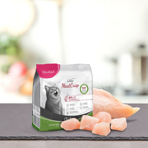 Vrečka PLATINUM MeatCrisp Sterilised Chicken Suha Hrana za Mačke s sivo mačko na sprednji strani stoji na pultu poleg kosov piščančjih prsi sveže meso na zamegljenem kuhinjskem ozadju.